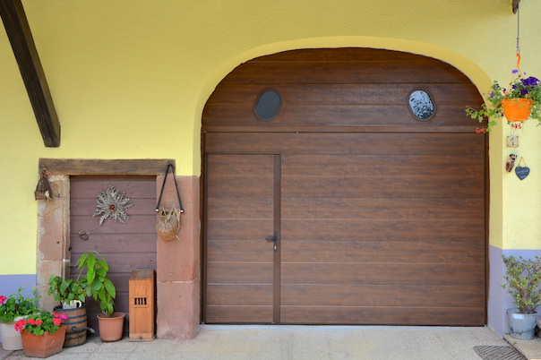 Porte de garage basculante chêne foncé avec portillon et un lambrequin avec deux hublots oblongs.